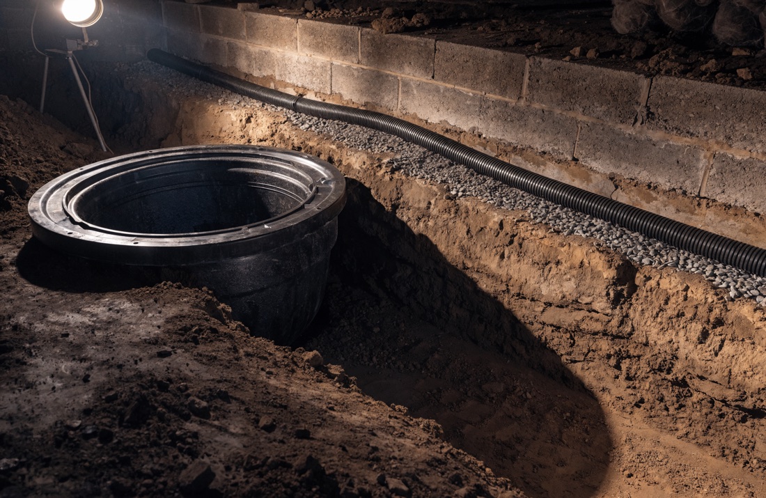 A sump basin set into a crawl space floor beside a gravel-filled perimeter trench with perforated drain pipe